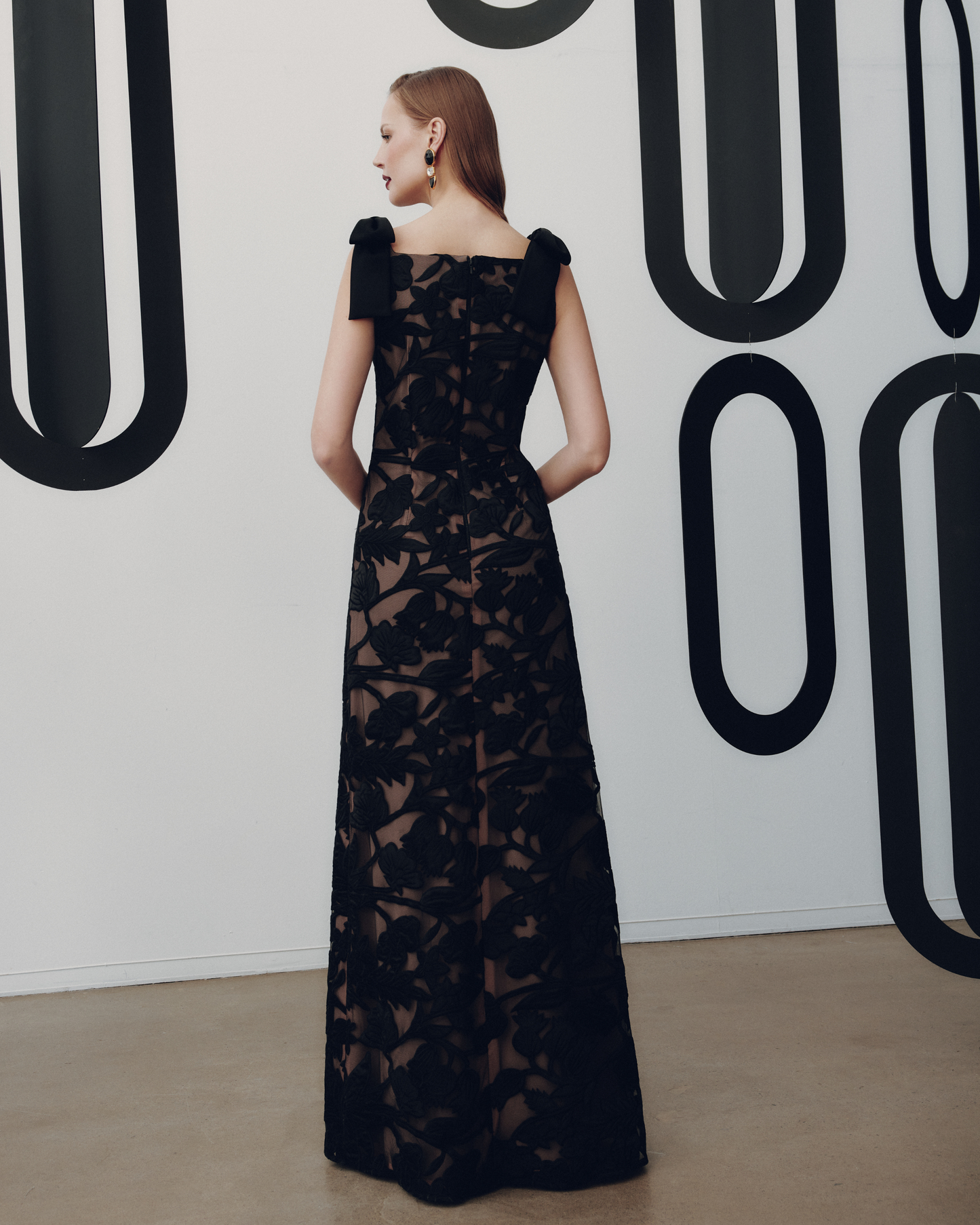 Woman wearing a black lace dress against a white wall with black abstract patterns.