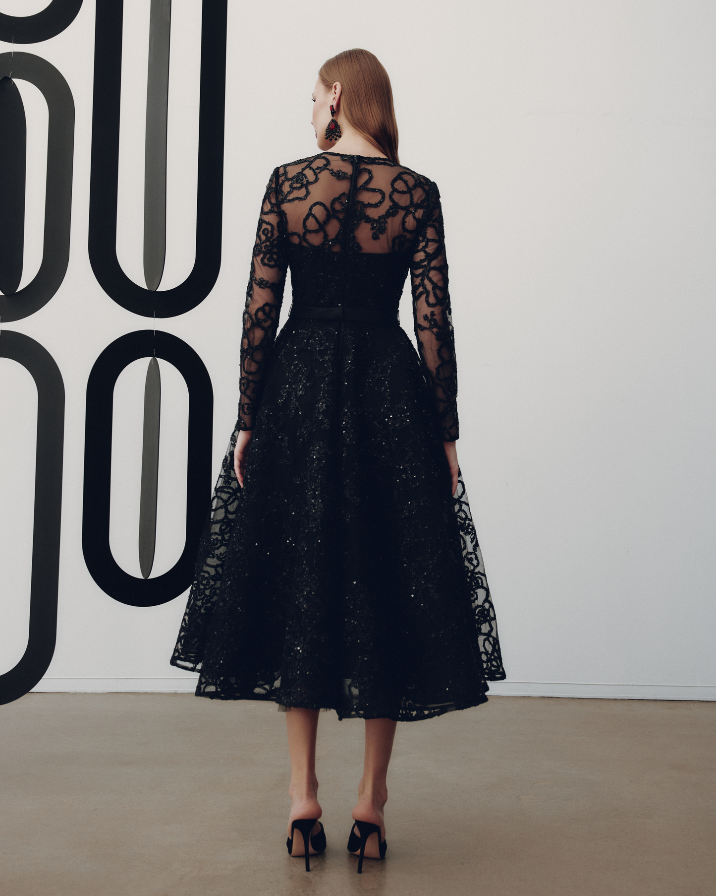 Woman wearing a black lace dress standing against a white wall with large black letters.