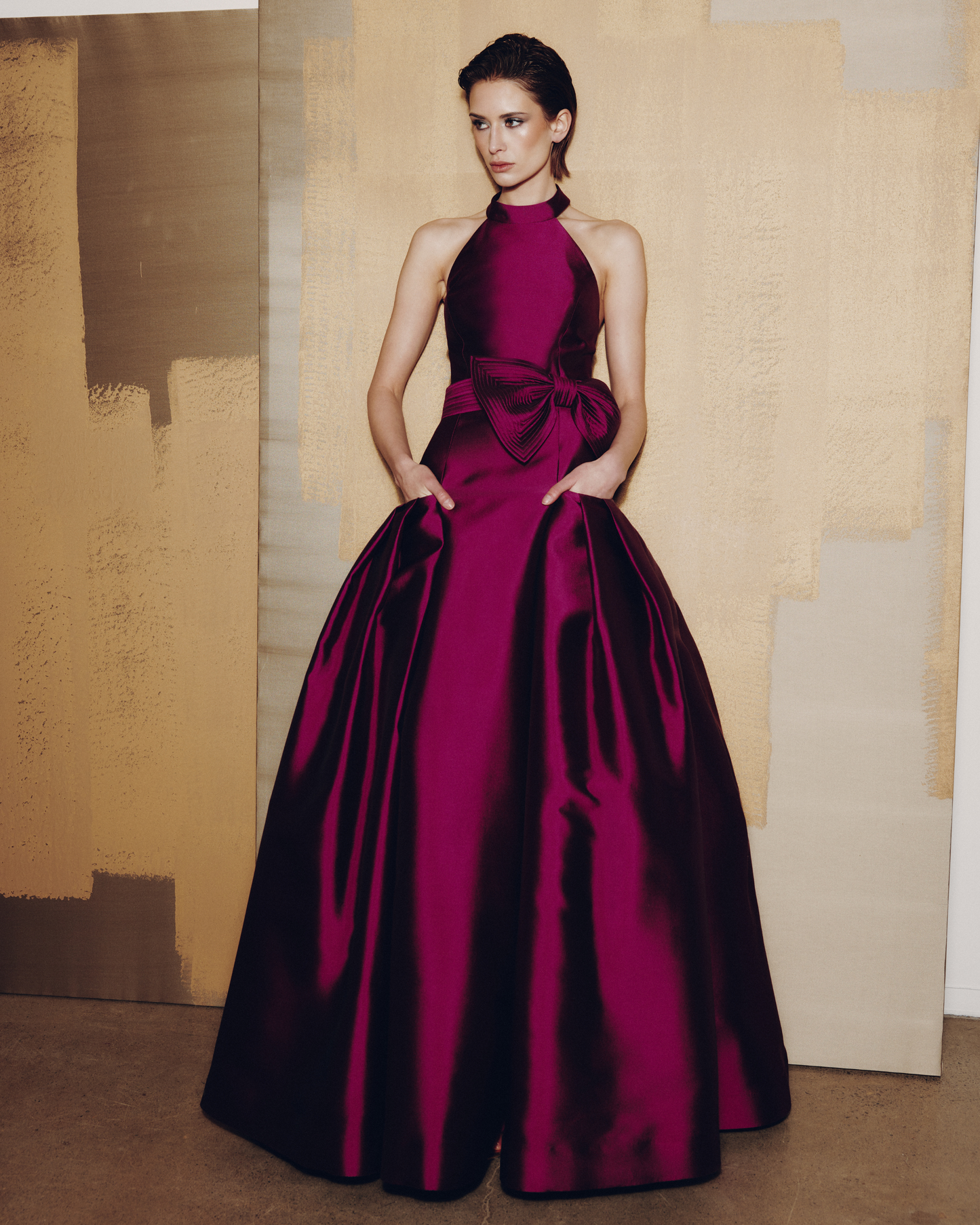 Woman wearing a burgundy evening gown with a large bow detail against a textured wall.