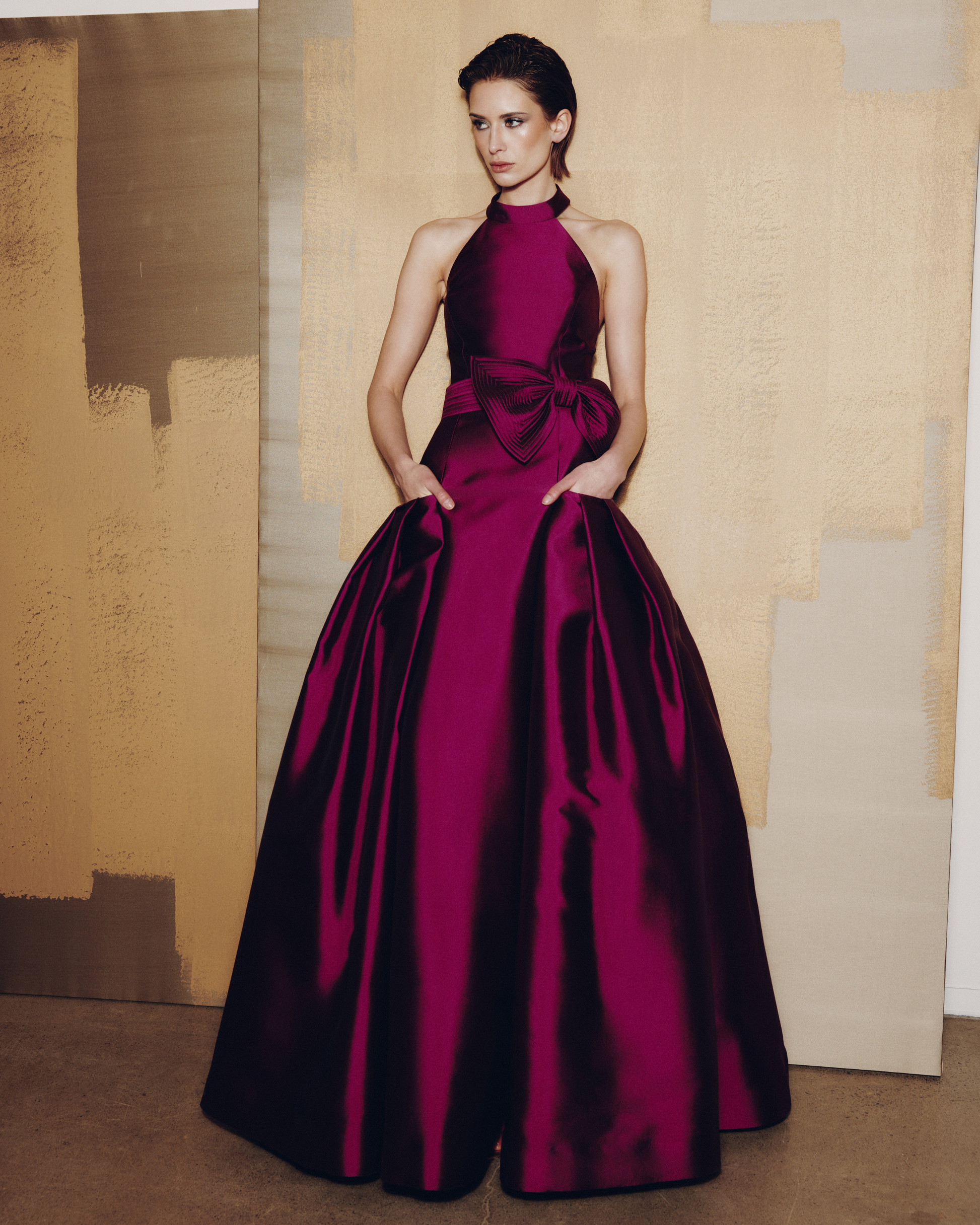 Woman wearing a burgundy evening gown with a large bow detail against a textured wall.