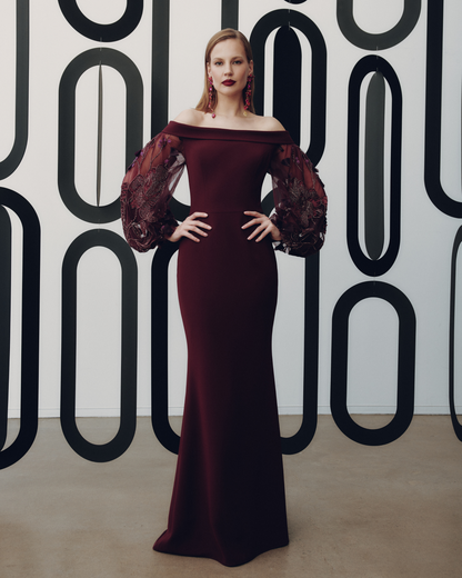 Woman in a burgundy off-shoulder gown with sheer sleeves against a patterned wall.