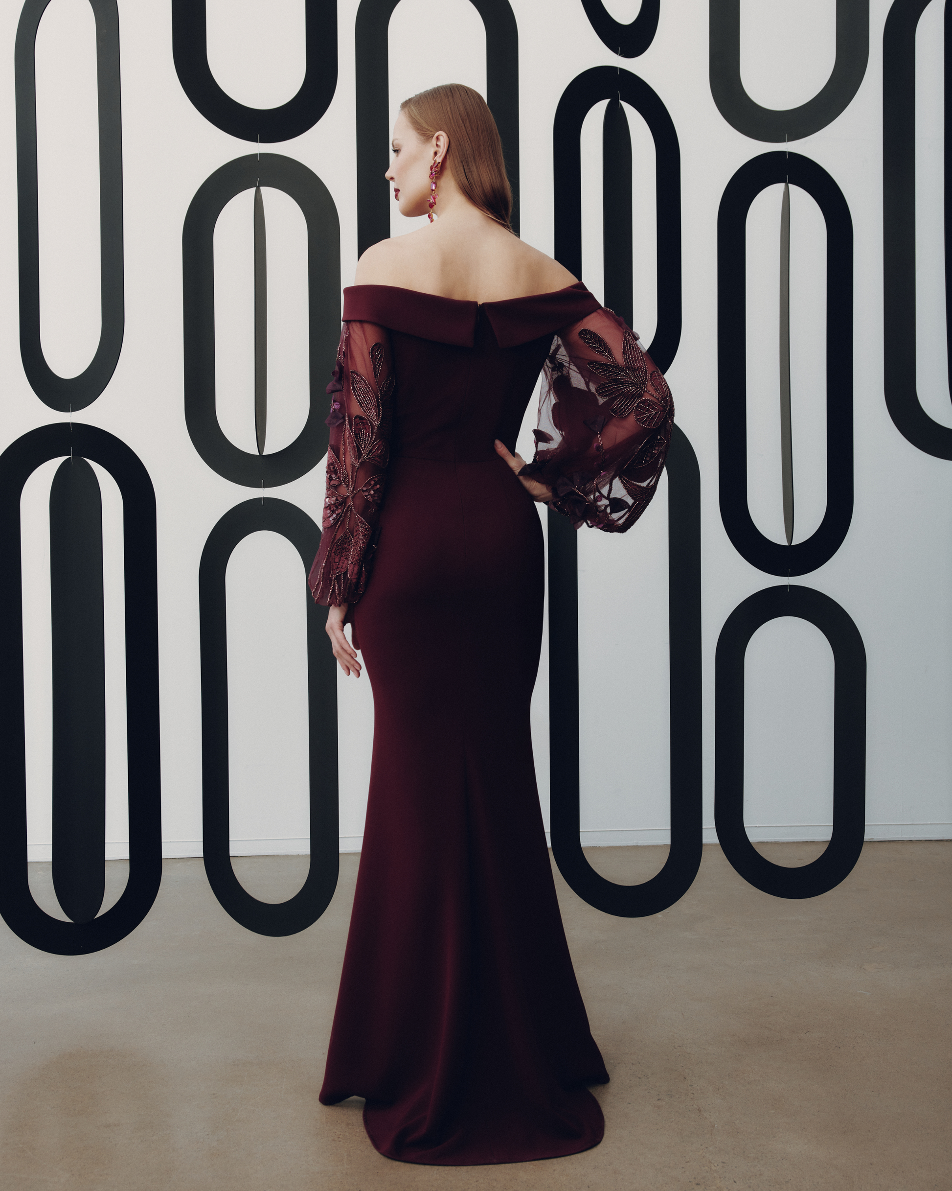 Woman in a burgundy off-shoulder gown against a geometric patterned wall.