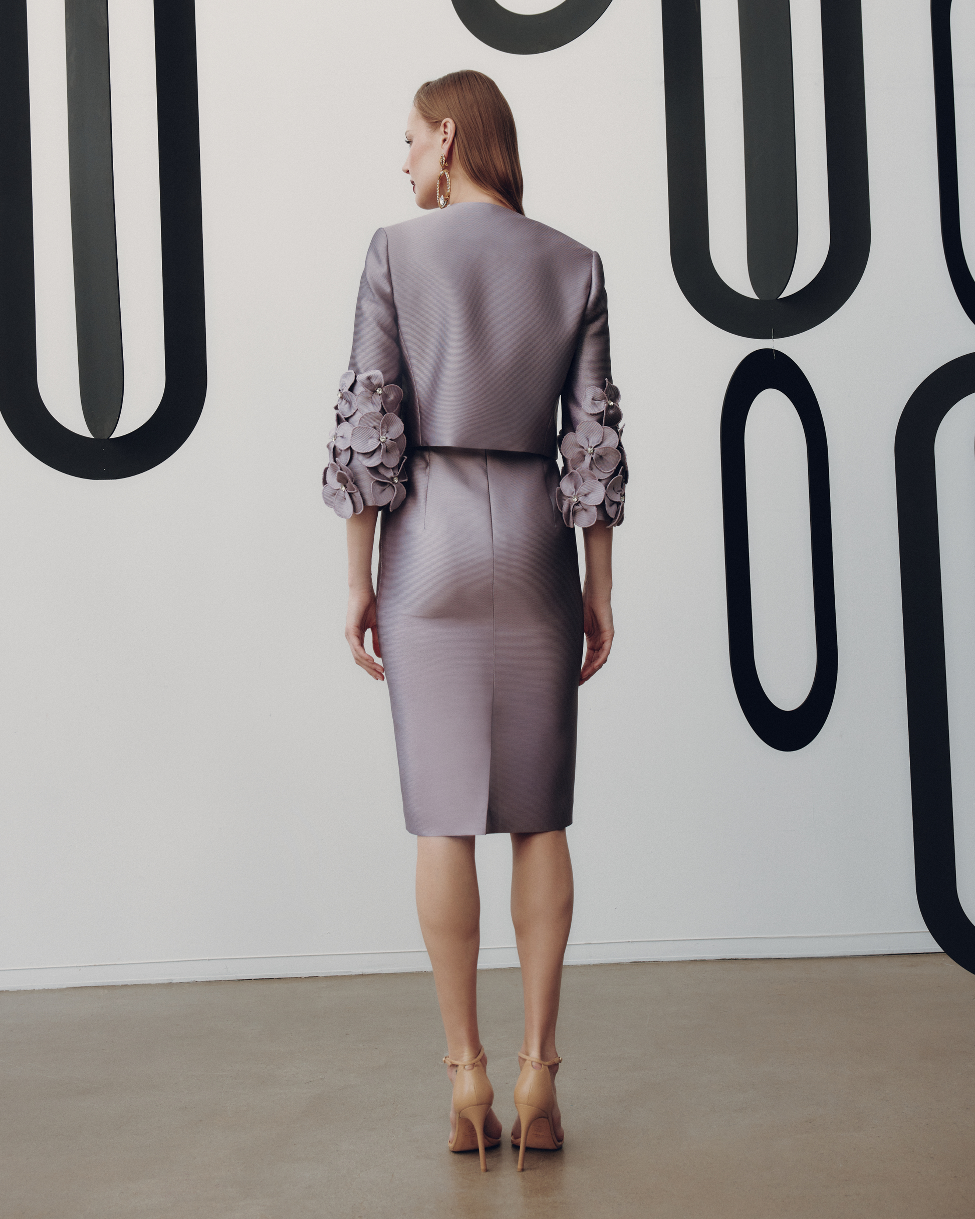 Woman in a gray suit with floral details standing against a white wall with black abstract patterns.