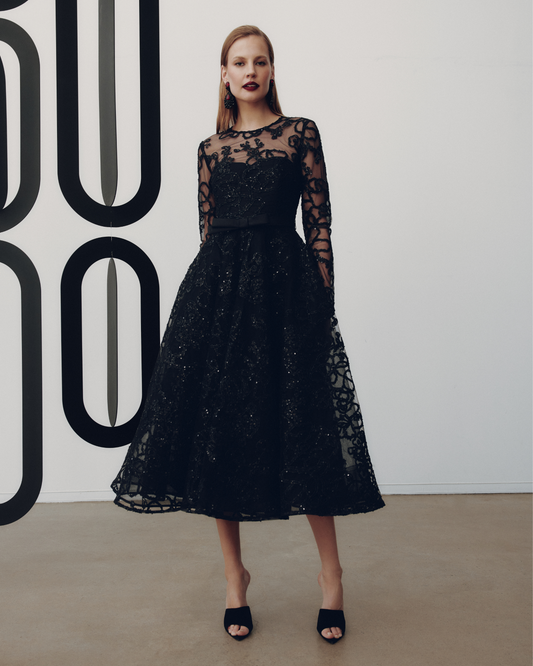 Woman wearing a black lace dress against a white wall with large black letters.