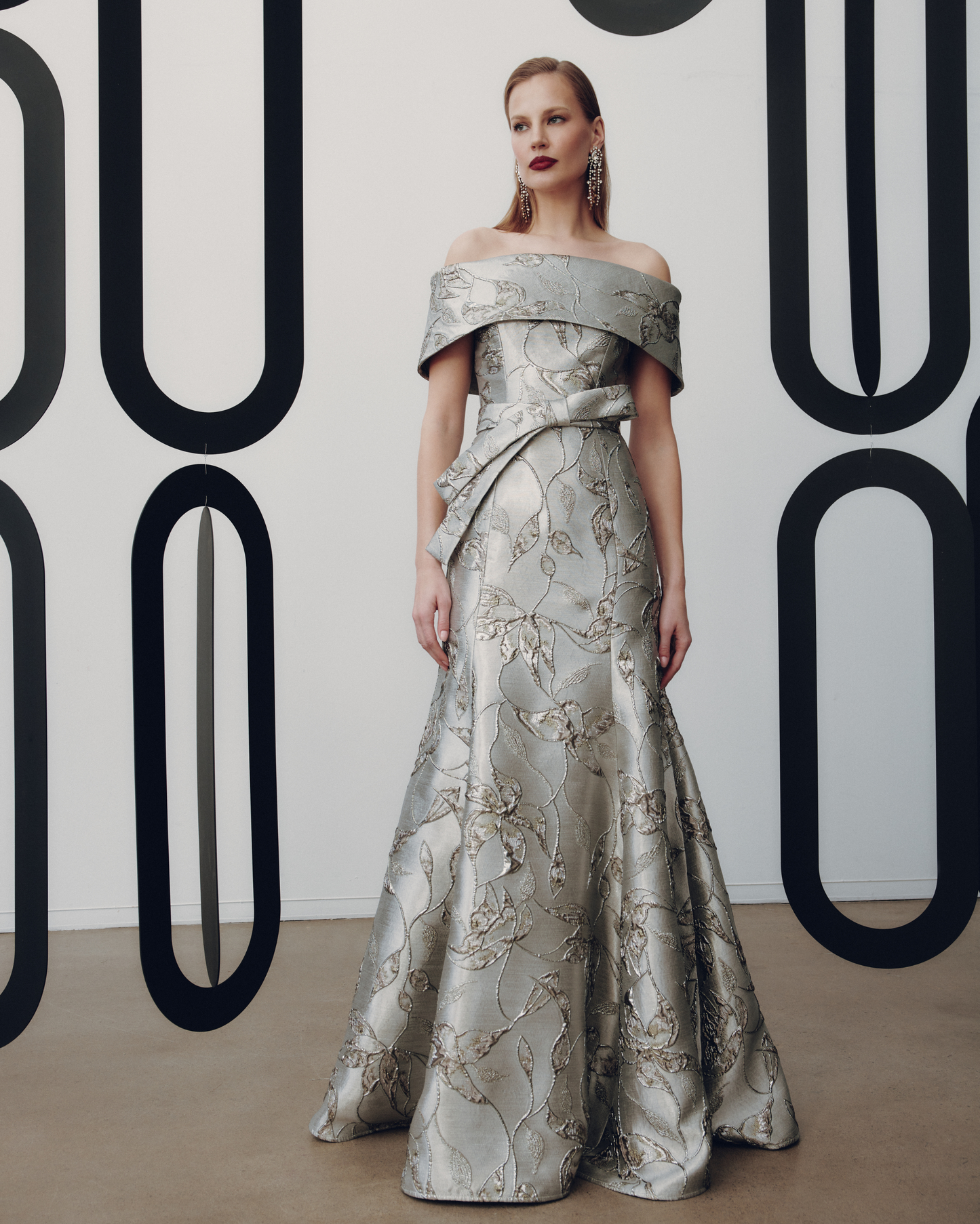 Woman in a silver off-shoulder gown standing against a decorative wall.