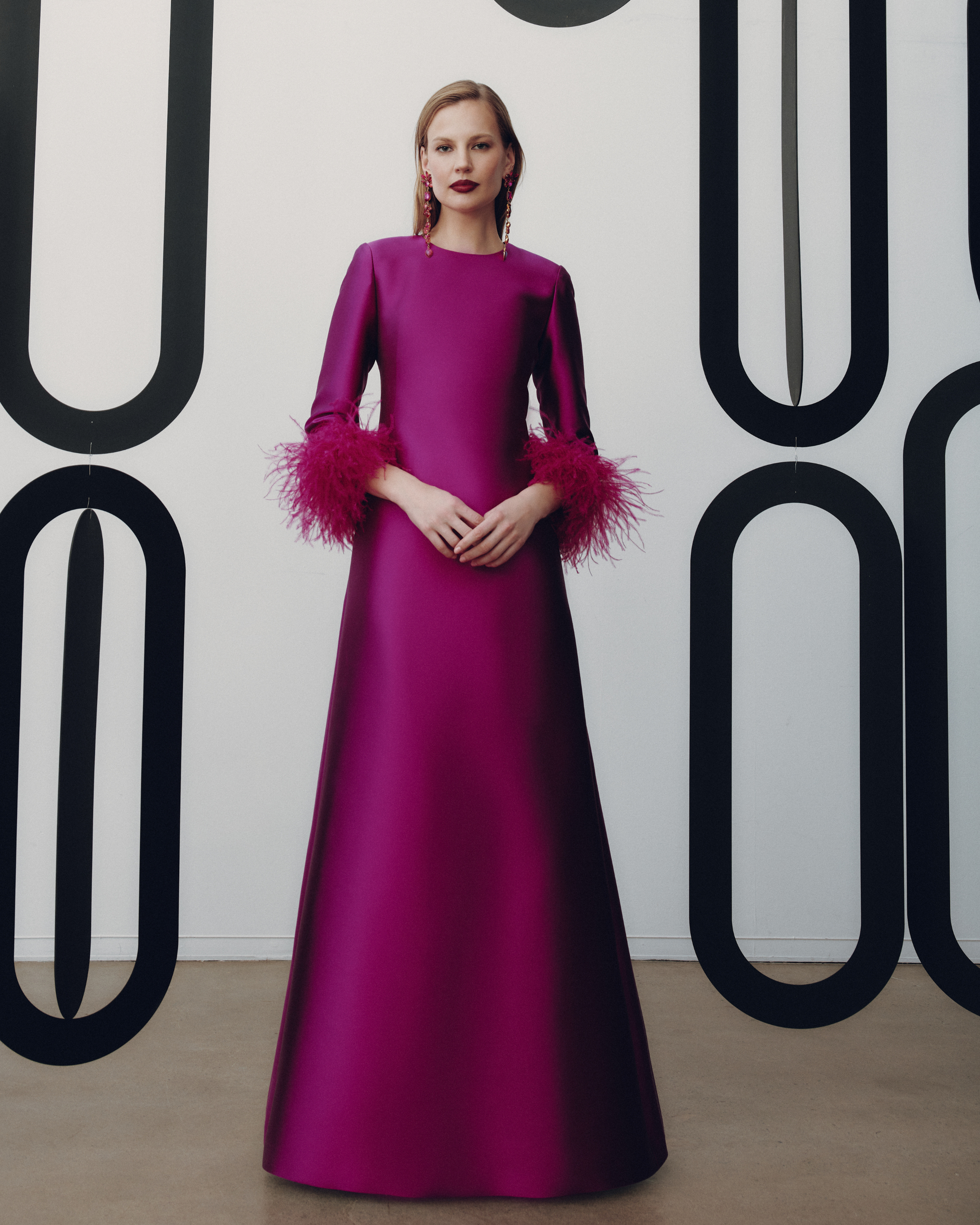 Woman wearing a magenta evening gown with feathered sleeves against a white background with black abstract patterns.